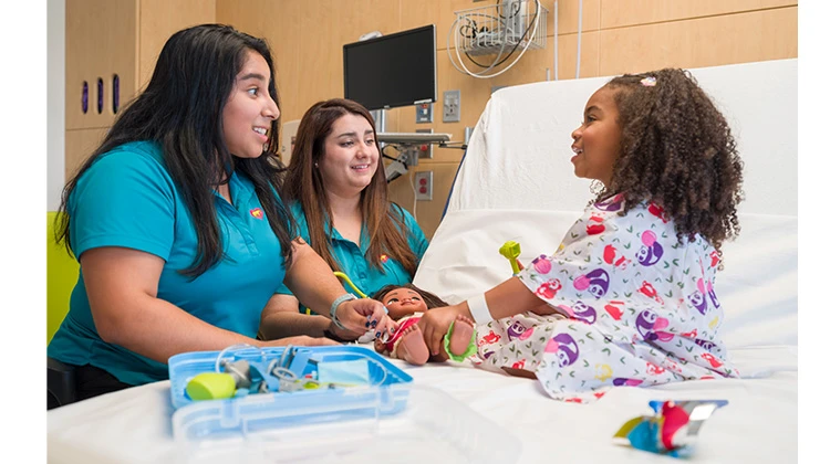 2 child life specialists playing games with girl at hospital bed.