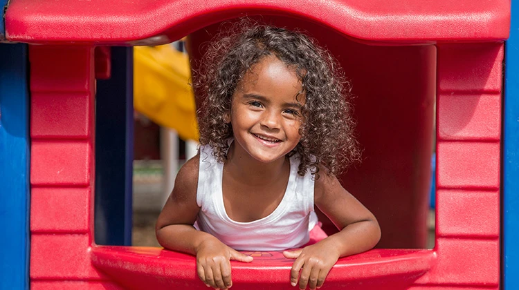 niña de cabello rizado feliz asomándose desde una casa de juego.